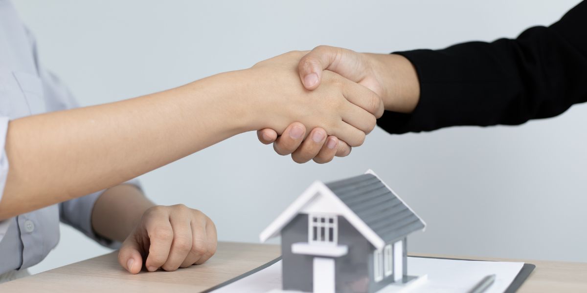 Two people shaking hands over a table with a small house model in the foreground.